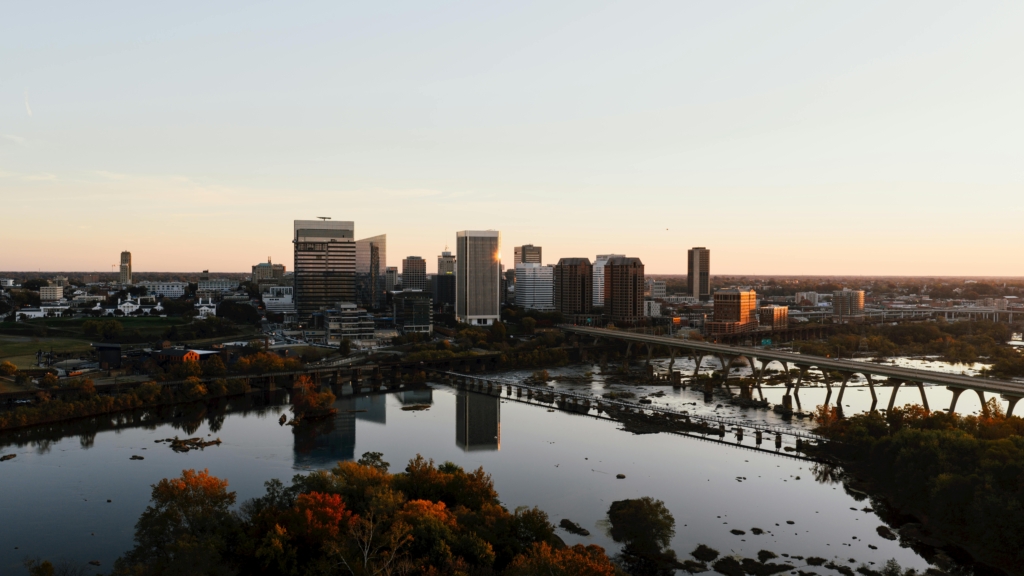 City skyline at sunset viewed from across a calm river with a bridge in the foreground.