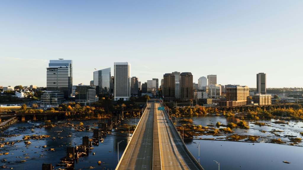 A city skyline at golden hour with a long bridge spanning a wide river in the foreground and modern glass office buildings in the background.