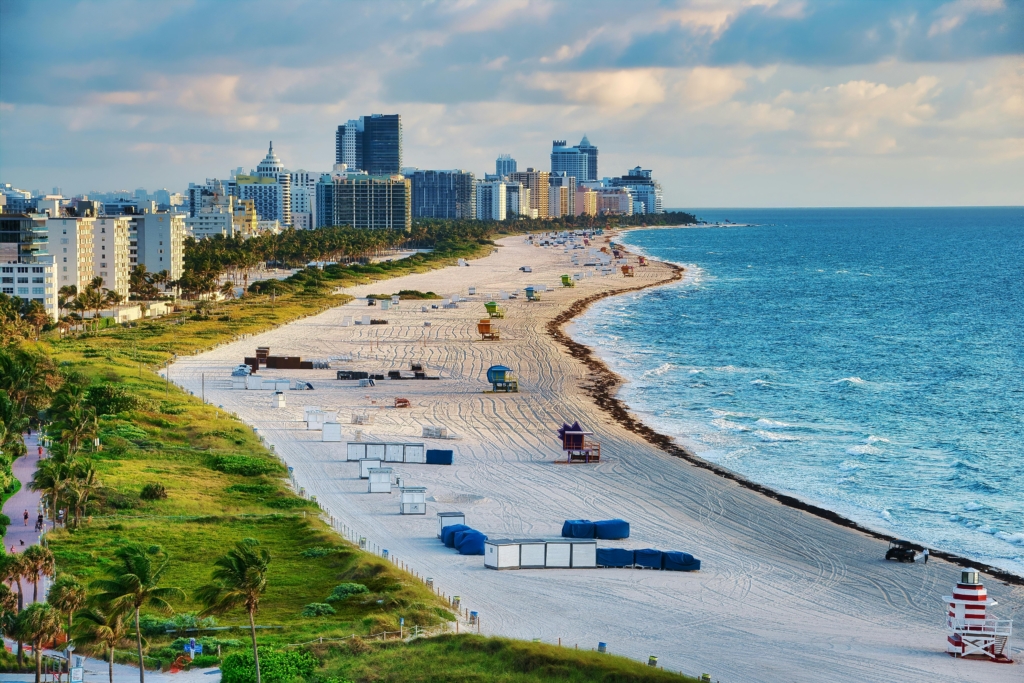 City skyline along a curved sandy beach with palm trees, high-rise hotels, and a blue sea under a partly cloudy sky awaiting visitors.